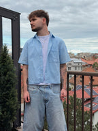 Man wearing a light blue shirt and jeans standing on a balcony with a cityscape background