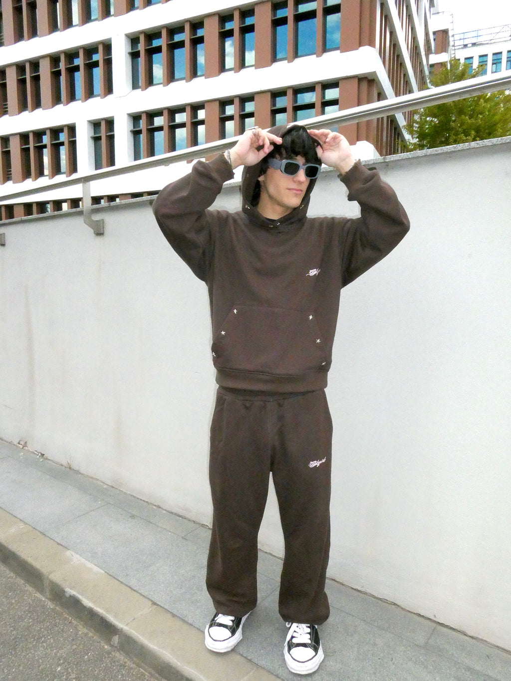 A person wearing a dark brown tracksuit with a matching hoodie and sweatpants, styled with black sunglasses and black-and-white sneakers, standing outdoors against a white wall and modern building.