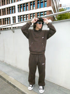 A person wearing a dark brown tracksuit with a matching hoodie and sweatpants, styled with black sunglasses and black-and-white sneakers, standing outdoors against a white wall and modern building.