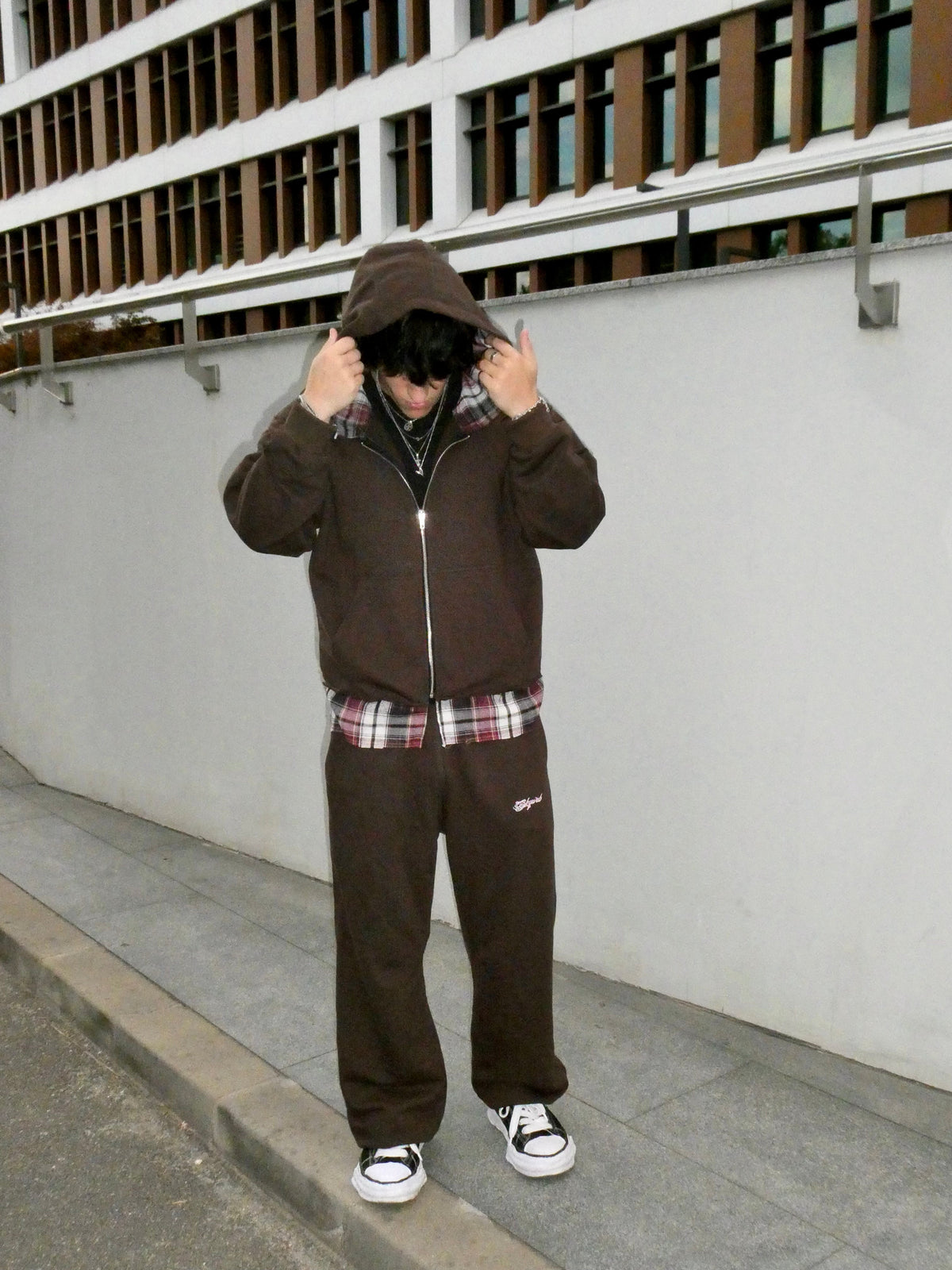 A person wearing a dark brown zip-up hoodie and matching sweatpants stands on a sidewalk in front of a white wall and modern building facade, holding the hood up with both hands.