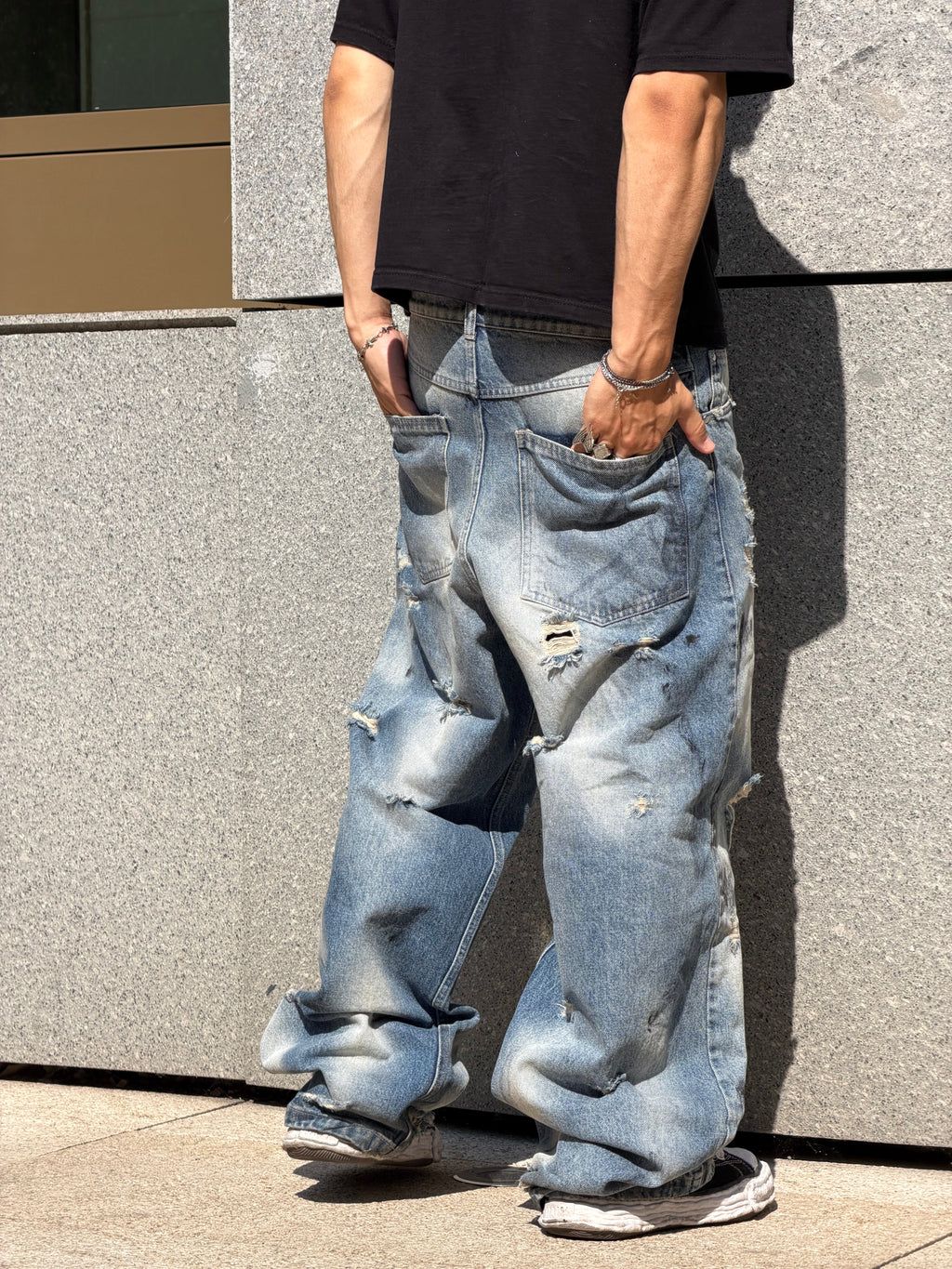 A person wearing extremely baggy, distressed light-wash blue jeans with ripped details and rolled cuffs, paired with a black t-shirt and sneakers, standing against a gray stone wall.