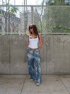 A woman stands outdoors against a concrete wall and chain-link fence, wearing a white ribbed tank top, distressed wide-leg blue jeans, and sunglasses.
