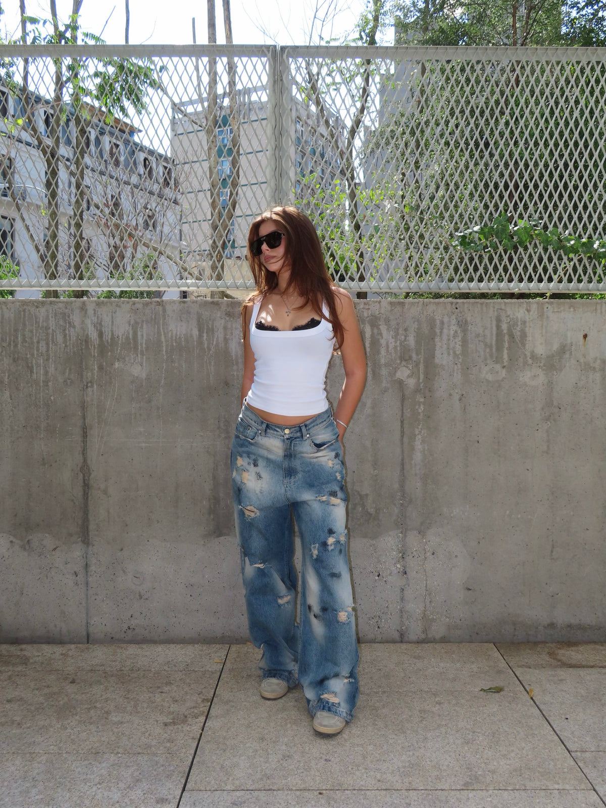 A woman stands outdoors against a concrete wall and chain-link fence, wearing a white ribbed tank top, distressed wide-leg blue jeans, and sunglasses.