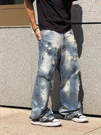 A person wearing distressed light blue wide-leg jeans with heavy fading and ripped details, paired with a black t-shirt and black sneakers, standing against a gray stone wall.