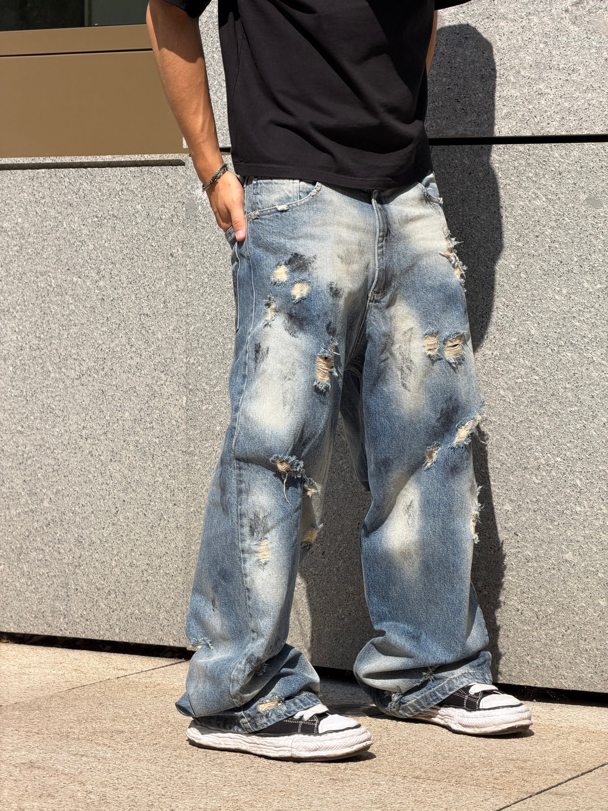 A person wearing distressed light blue wide-leg jeans with heavy fading and ripped details, paired with a black t-shirt and black sneakers, standing against a gray stone wall.