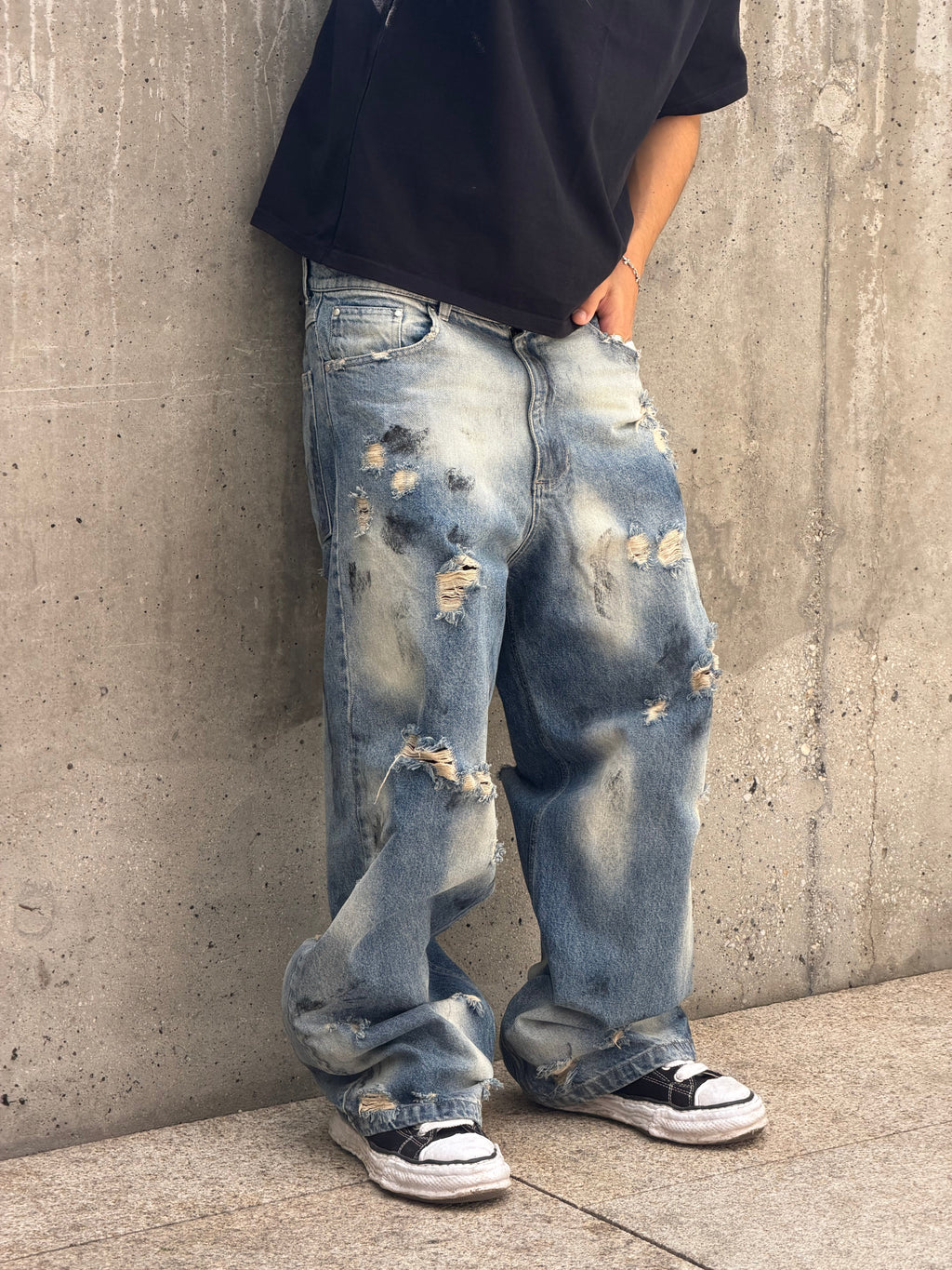 A person wearing oversized light-wash distressed jeans with heavy fading and ripped details, paired with a black t-shirt and black-and-white sneakers, standing against a concrete wall.