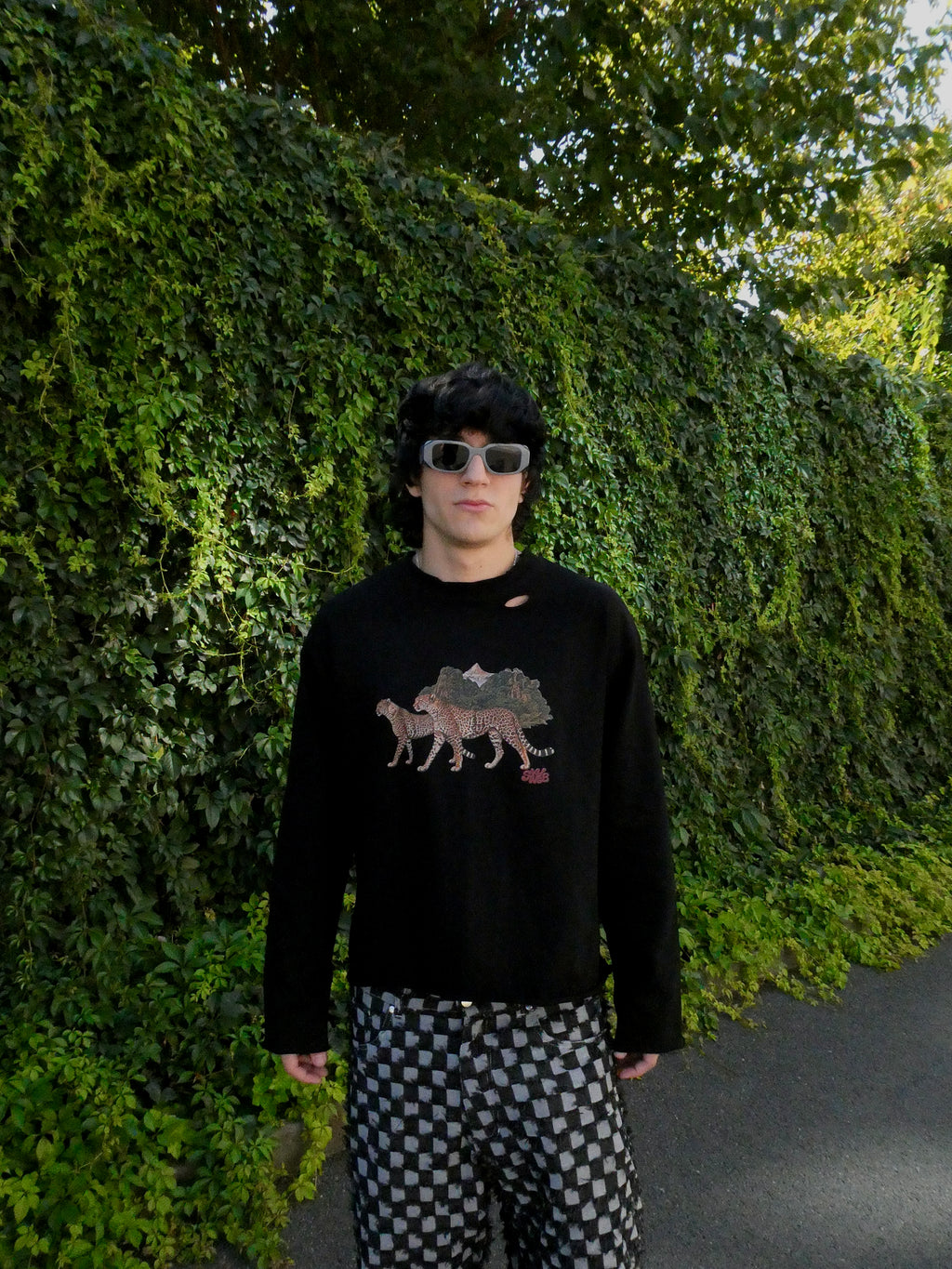 A person standing outdoors in front of a dense green ivy-covered wall, wearing a black crewneck sweatshirt with a graphic of three camels and sunglasses, paired with black and white checkered pants.