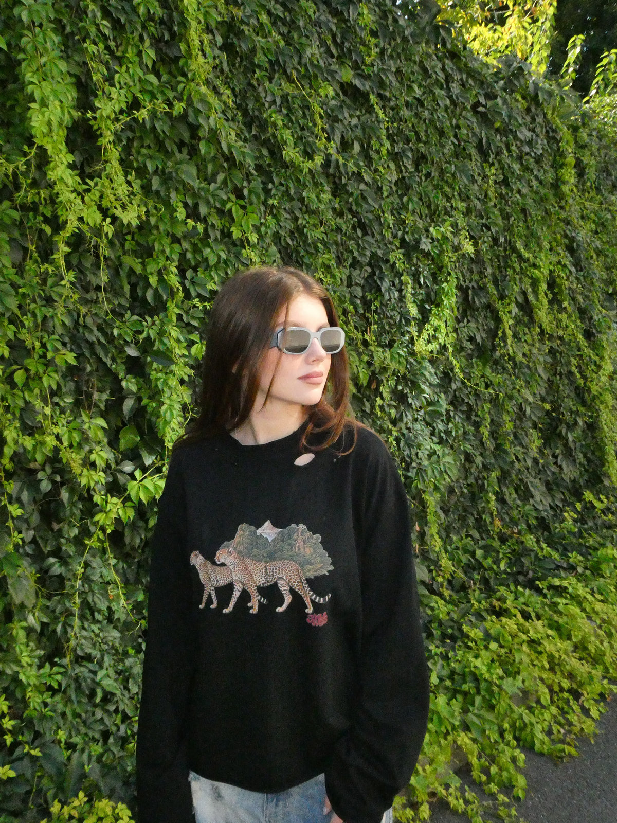 A woman wearing a black crewneck sweatshirt with a detailed camel graphic on the front, paired with light blue jeans and sunglasses, standing in front of a dense green ivy wall.