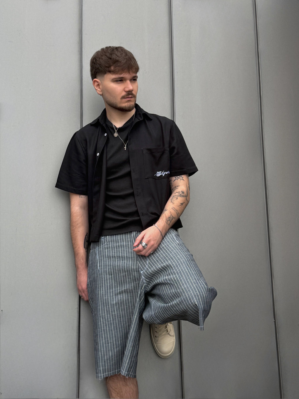 Man wearing a black shirt and plaid shorts leaning against a gray wall.