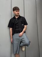 Man wearing a black shirt and plaid shorts leaning against a gray wall.