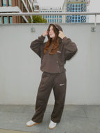A woman wearing a matching brown hooded sweatshirt and sweatpants set with small white embroidered logos, posing outdoors against a light gray wall.