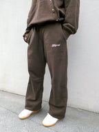 A person wearing loose brown sweatpants and a matching brown sweatshirt, standing against a light gray wall. The sweatpants have a small white script logo on the left thigh and are paired with white slip-on shoes.