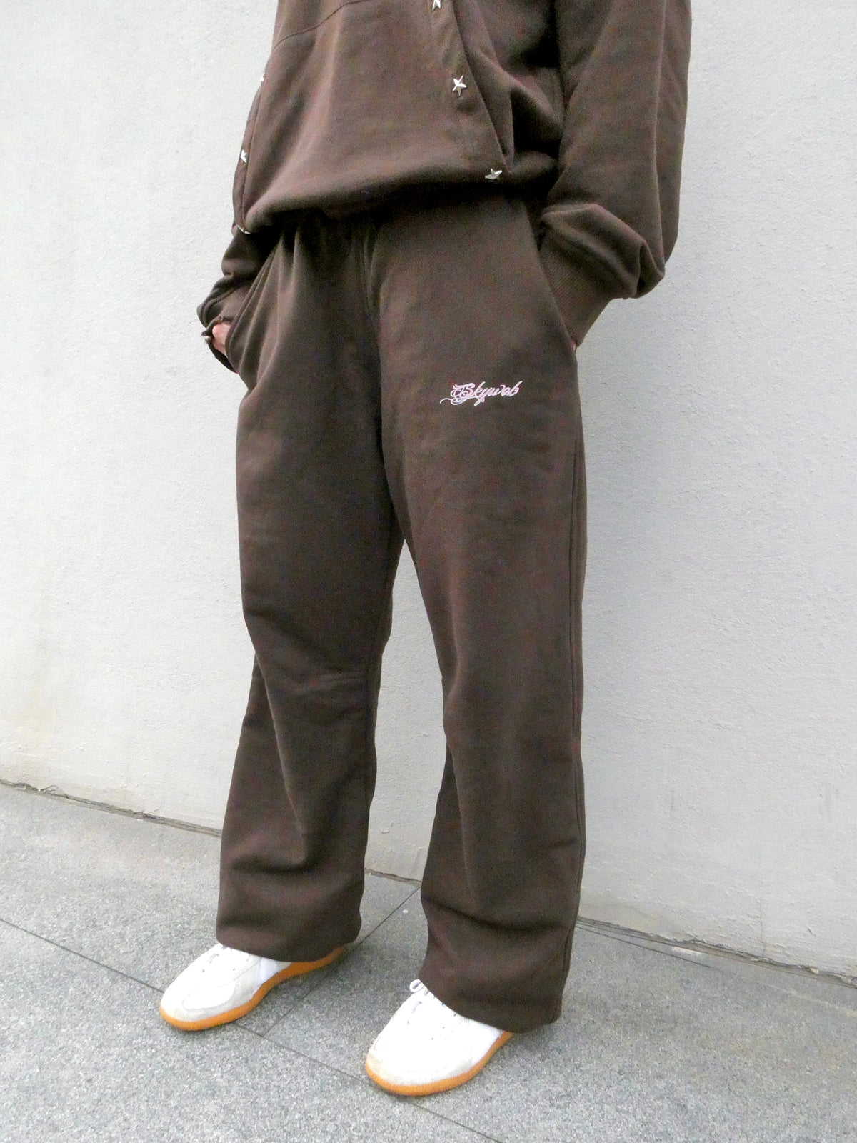 A person wearing loose brown sweatpants and a matching brown sweatshirt, standing against a light gray wall. The sweatpants have a small white script logo on the left thigh and are paired with white slip-on shoes.