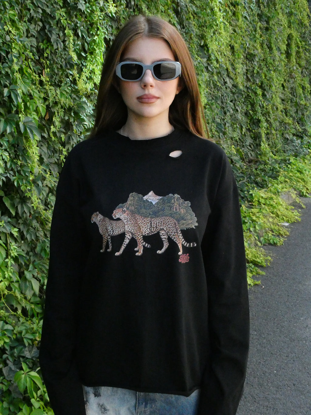 A woman wearing a black crewneck sweatshirt with a distressed hole in the chest and a graphic of two cheetahs walking in front of mountains, standing outdoors against a green ivy-covered wall.