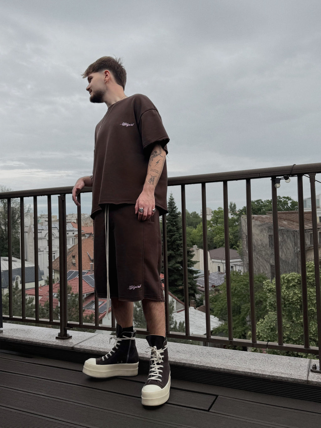Man wearing a brown outfit standing on a balcony with a cityscape background
