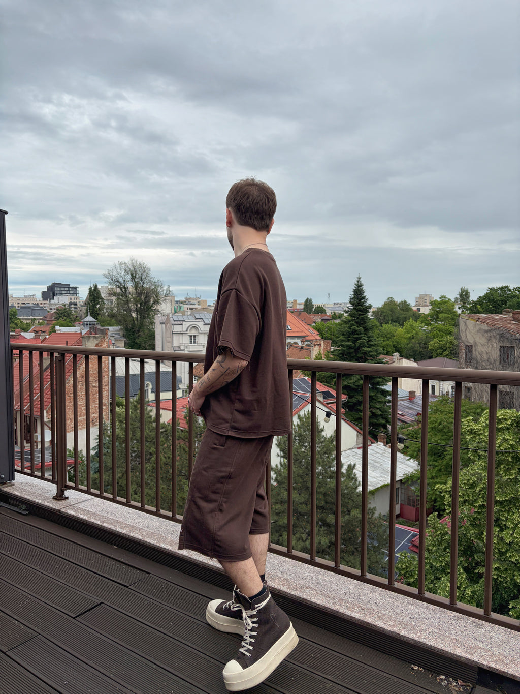 Person standing on a balcony overlooking a cityscape with green trees and rooftops.