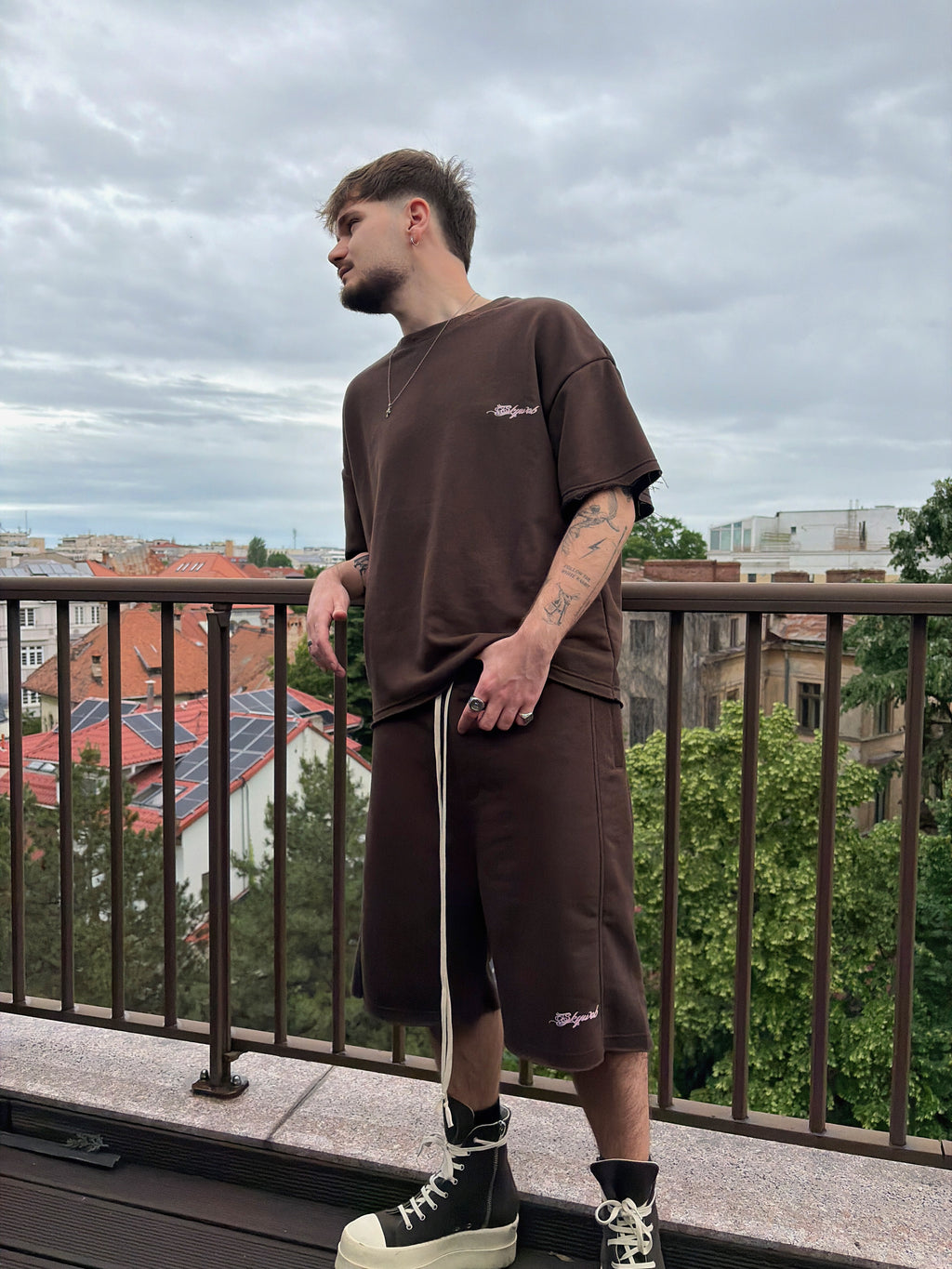 Man wearing a brown outfit standing on a balcony with a cityscape in the background
