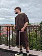 Man wearing a brown outfit standing on a balcony with a cityscape in the background