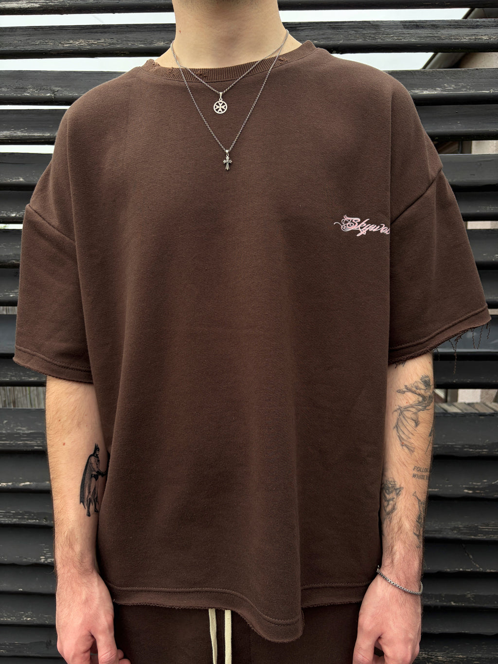 Person wearing a brown t-shirt with a small logo, standing against a metal slat background.