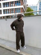 A person wearing a dark brown hooded sweatshirt and matching sweatpants stands on a sidewalk next to a gray concrete wall, with modern buildings in the background.