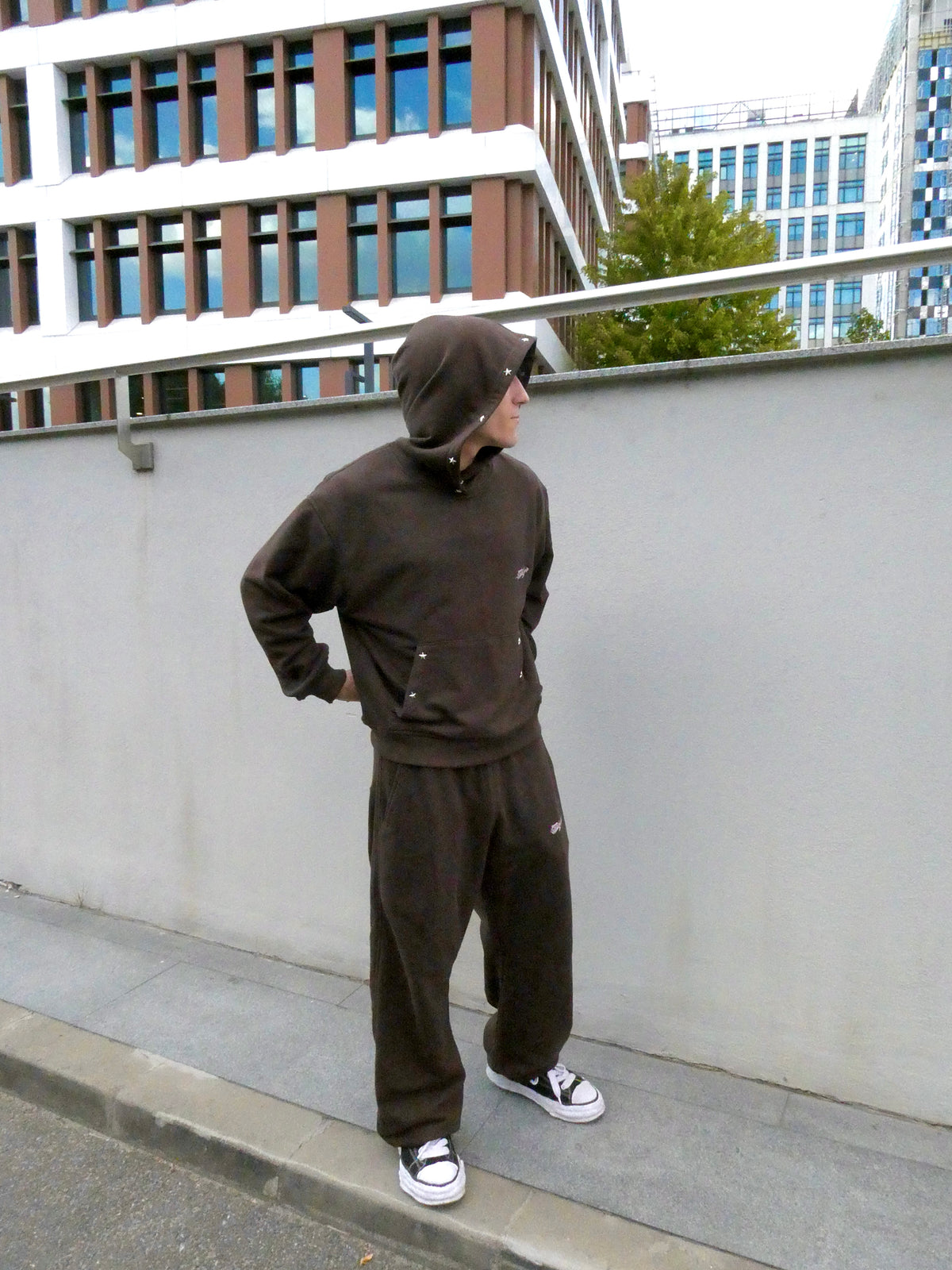 A person wearing a dark brown hooded sweatshirt and matching sweatpants stands on a sidewalk next to a gray concrete wall, with modern buildings in the background.
