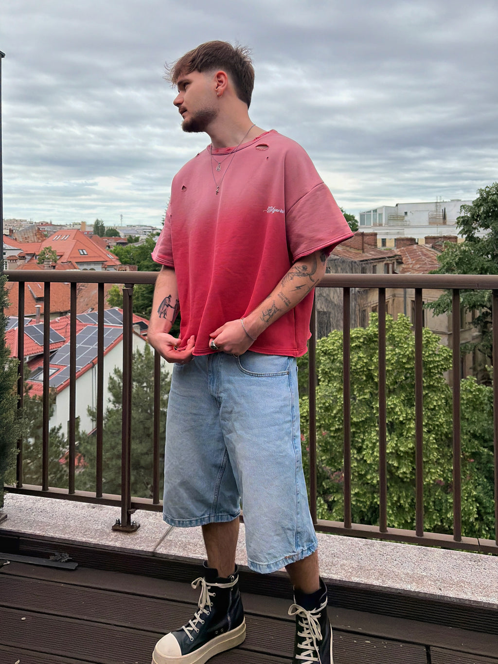 Person wearing a red shirt and light blue shorts standing on a balcony with a cityscape background