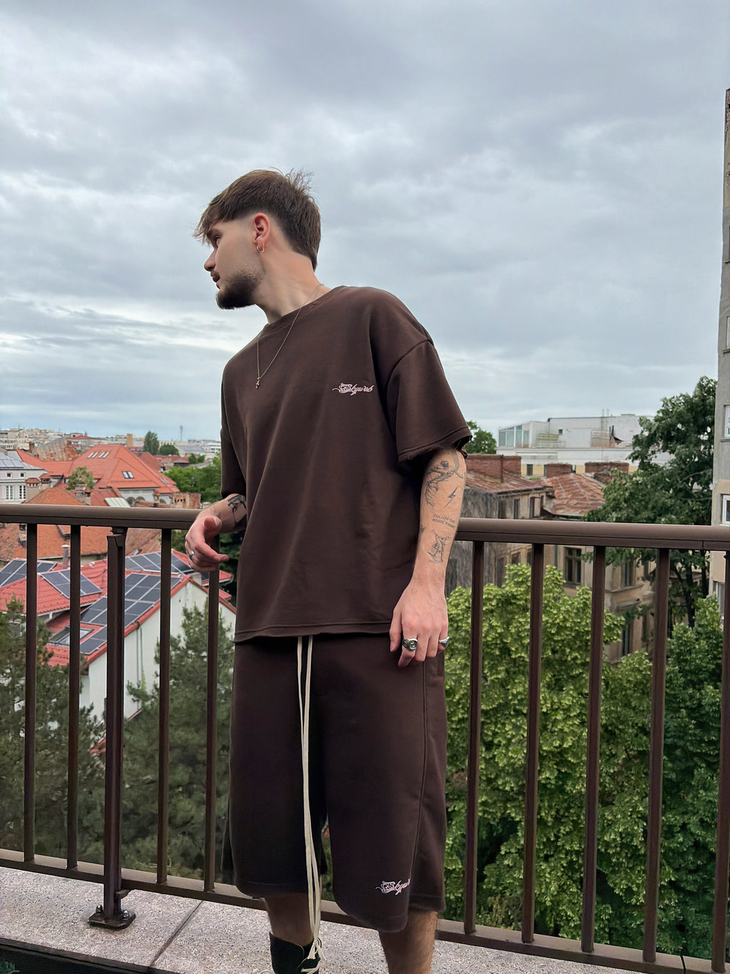 Man wearing a brown t-shirt and shorts standing on a balcony with a cityscape in the background