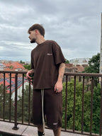 Man wearing a brown t-shirt and shorts standing on a balcony with a cityscape in the background