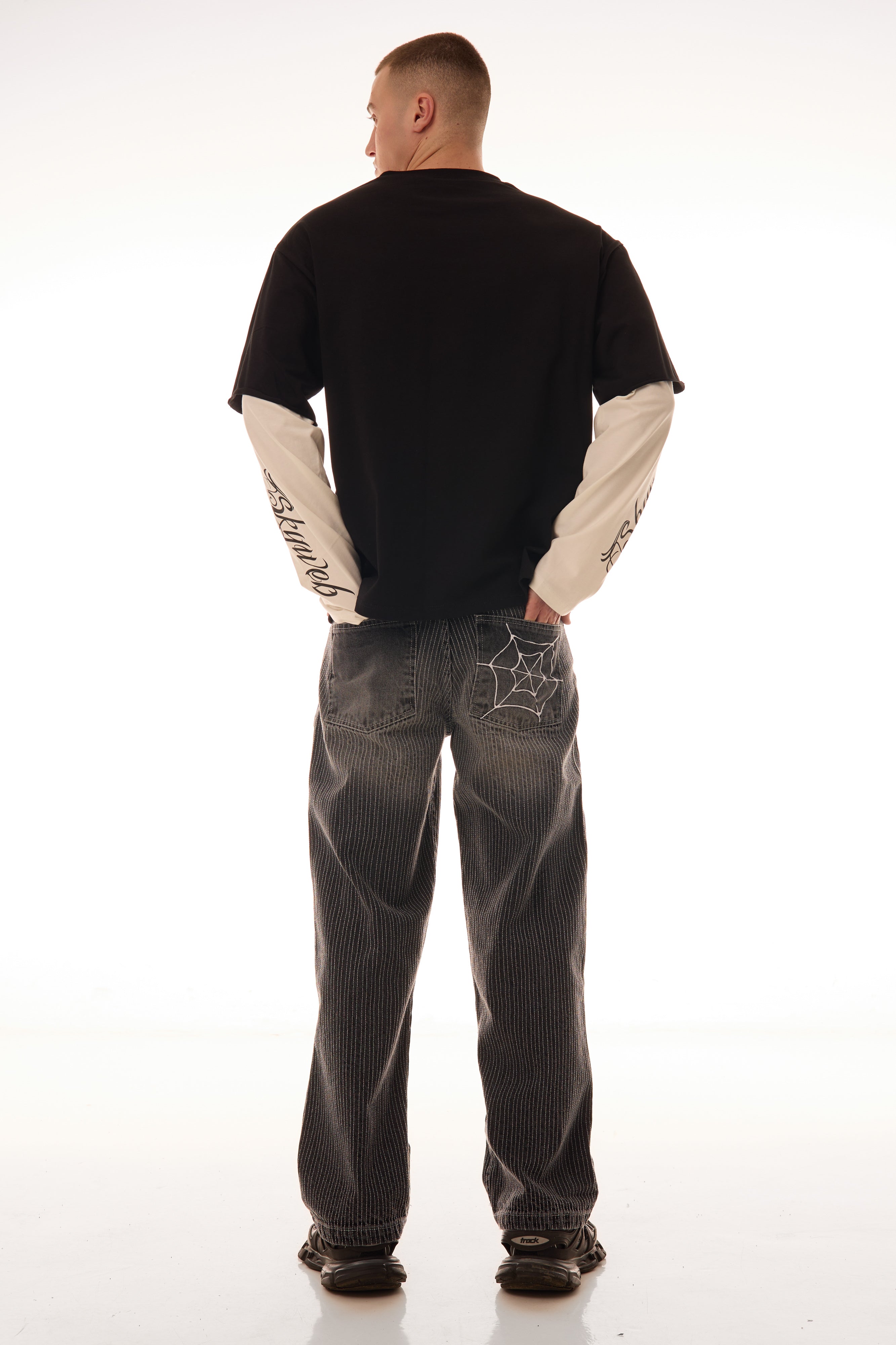 Model văzut din spate purtând blugii Axis Denim gri, wide fit, cu tighele verticale albe și broderie pe buzunarul spate.
