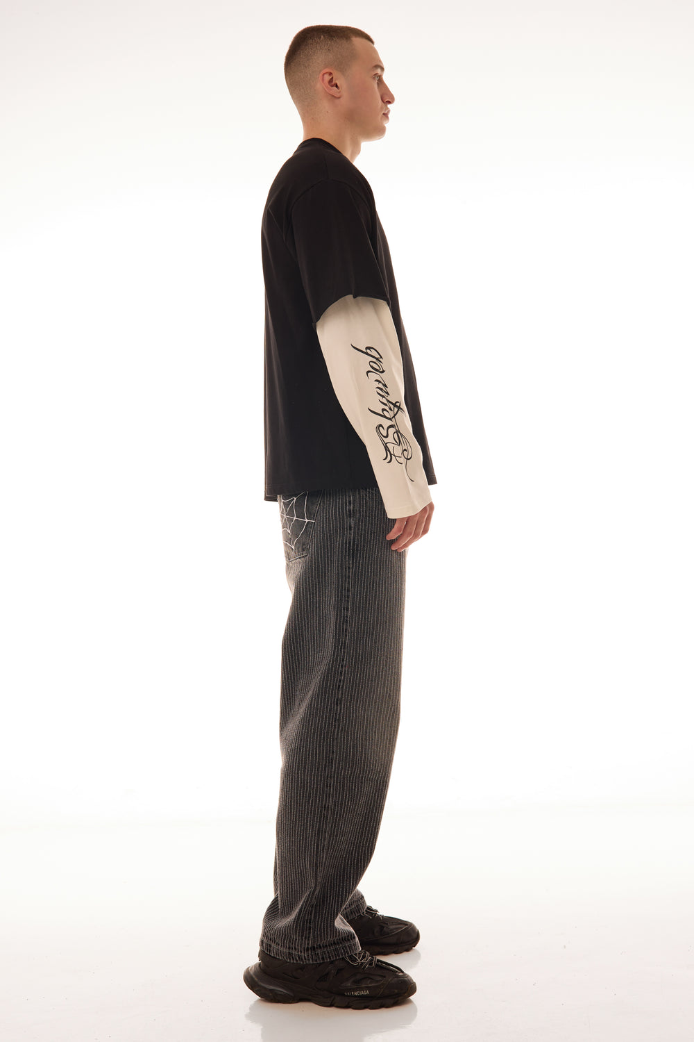 Model văzut din lateral purtând tricoul DUAL LONG TEE Skyweb, tricou negru cu mâneci albe layered și print gotic pe mâneci.