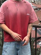 Person wearing a red t-shirt with a logo and blue jeans, standing on a balcony with buildings in the background.